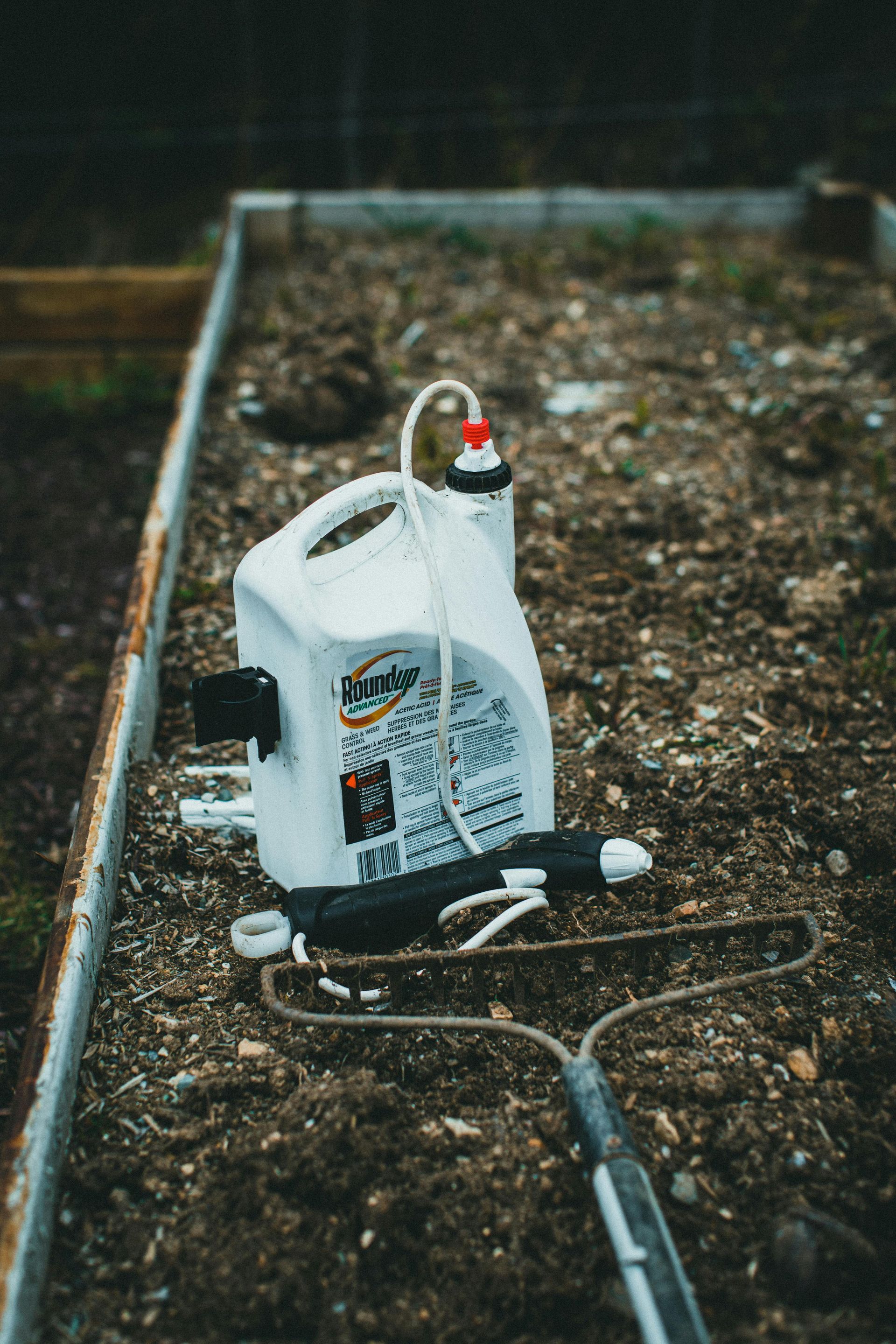 White sprayer and rake in a garden bed with dark soil.