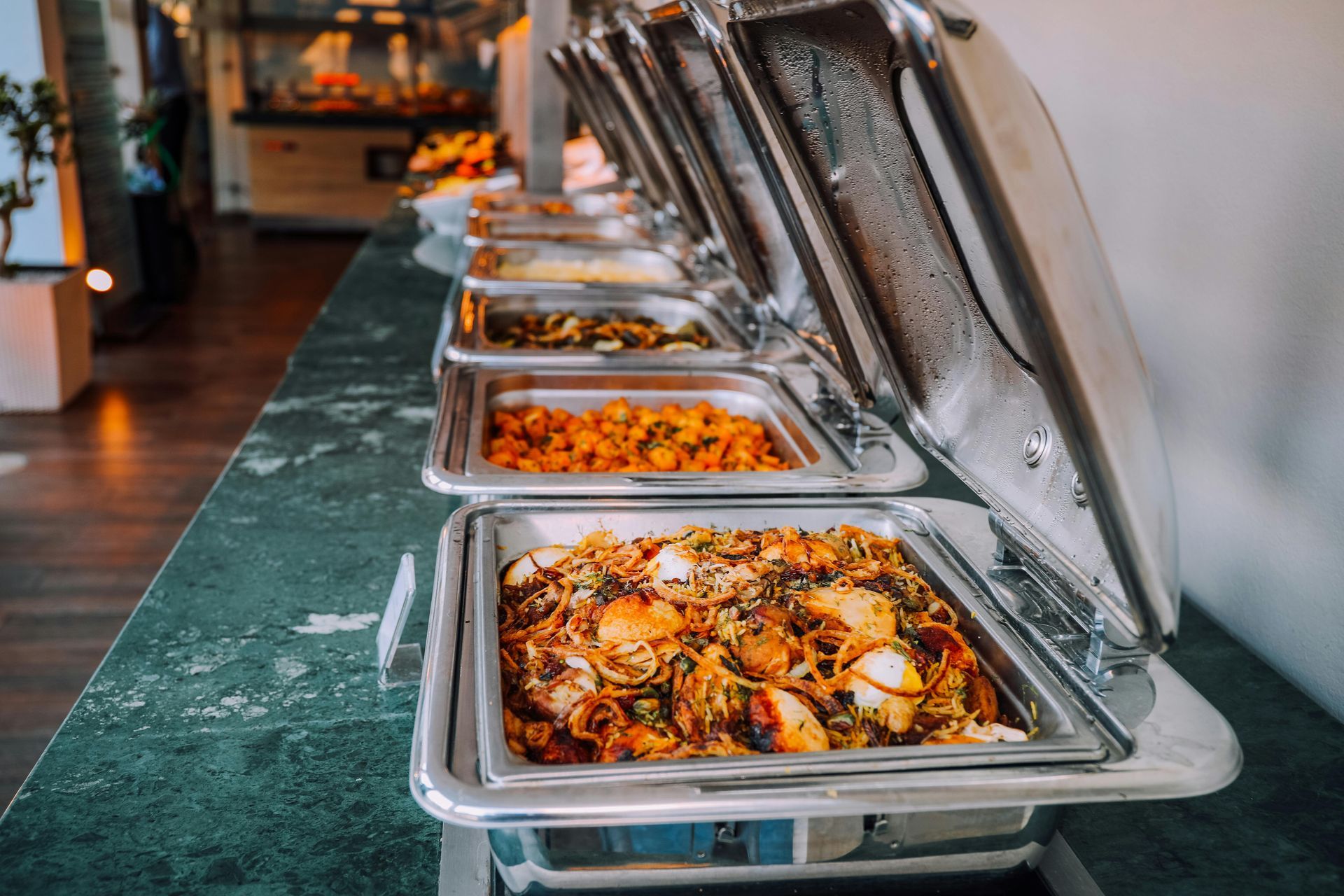 Buffet with various food options in stainless steel warming trays, set on a green countertop.