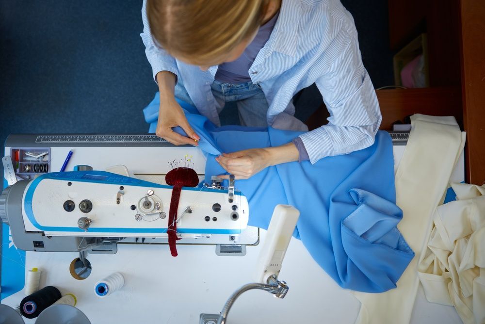 Person sewing blue fabric on a machine with visible red thread in a workroom