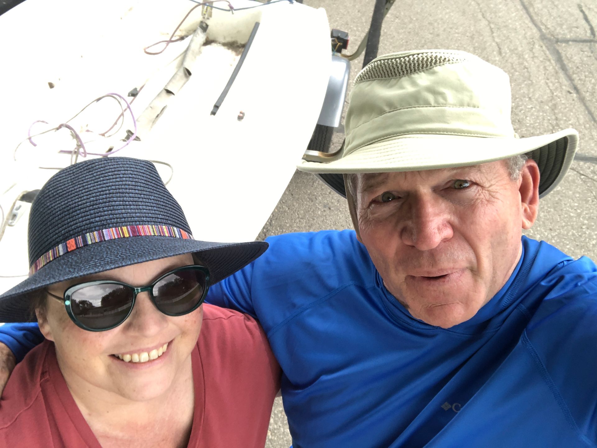 Two smiling people in sun hats and sunglasses pose outdoors near a boat.