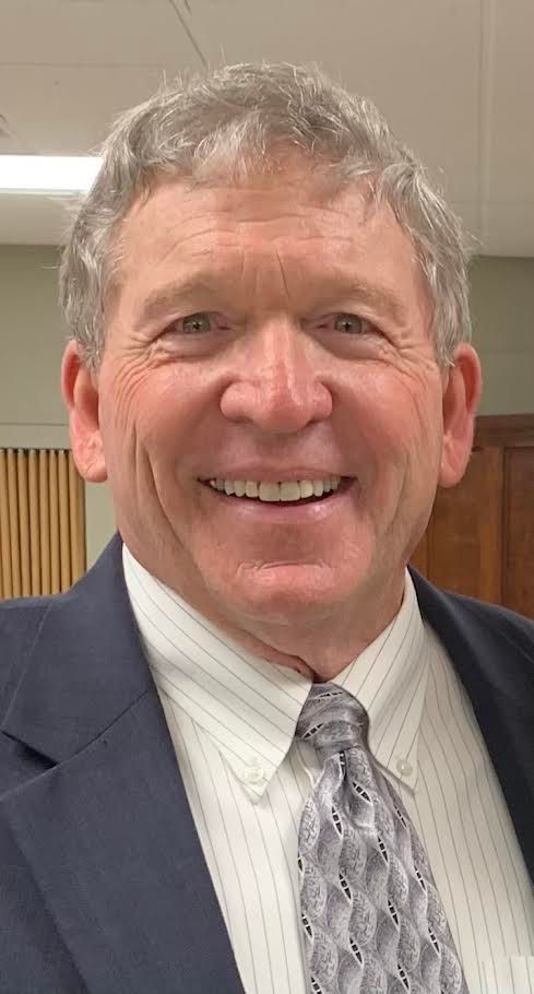 Smiling man in a suit and tie standing indoors against a wood-paneled wall