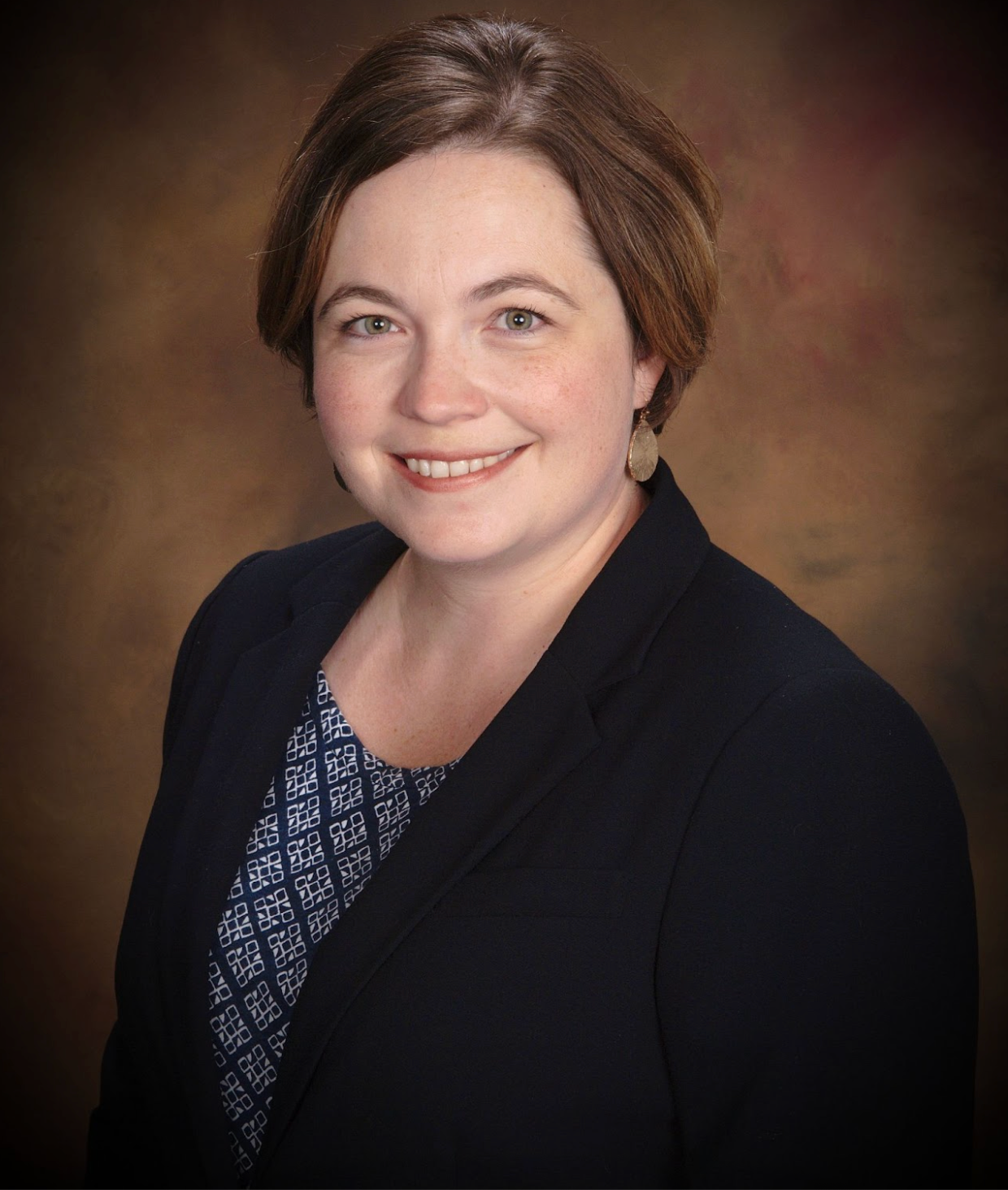 Studio portrait of a smiling person in a dark blazer with a patterned top on a brown background