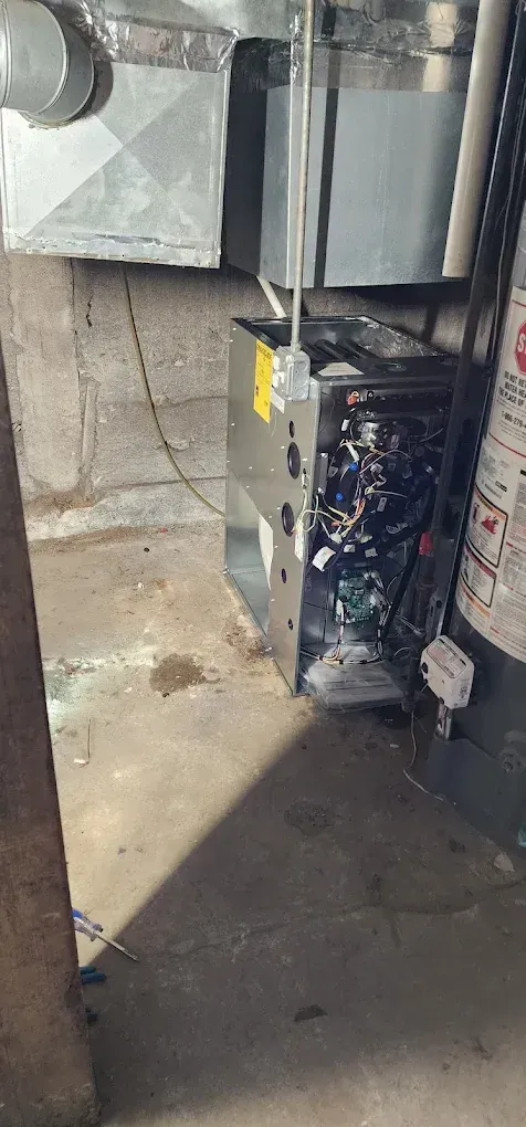 A furnace sits in a basement. Metal ductwork is above. A water heater is nearby. Concrete floor.