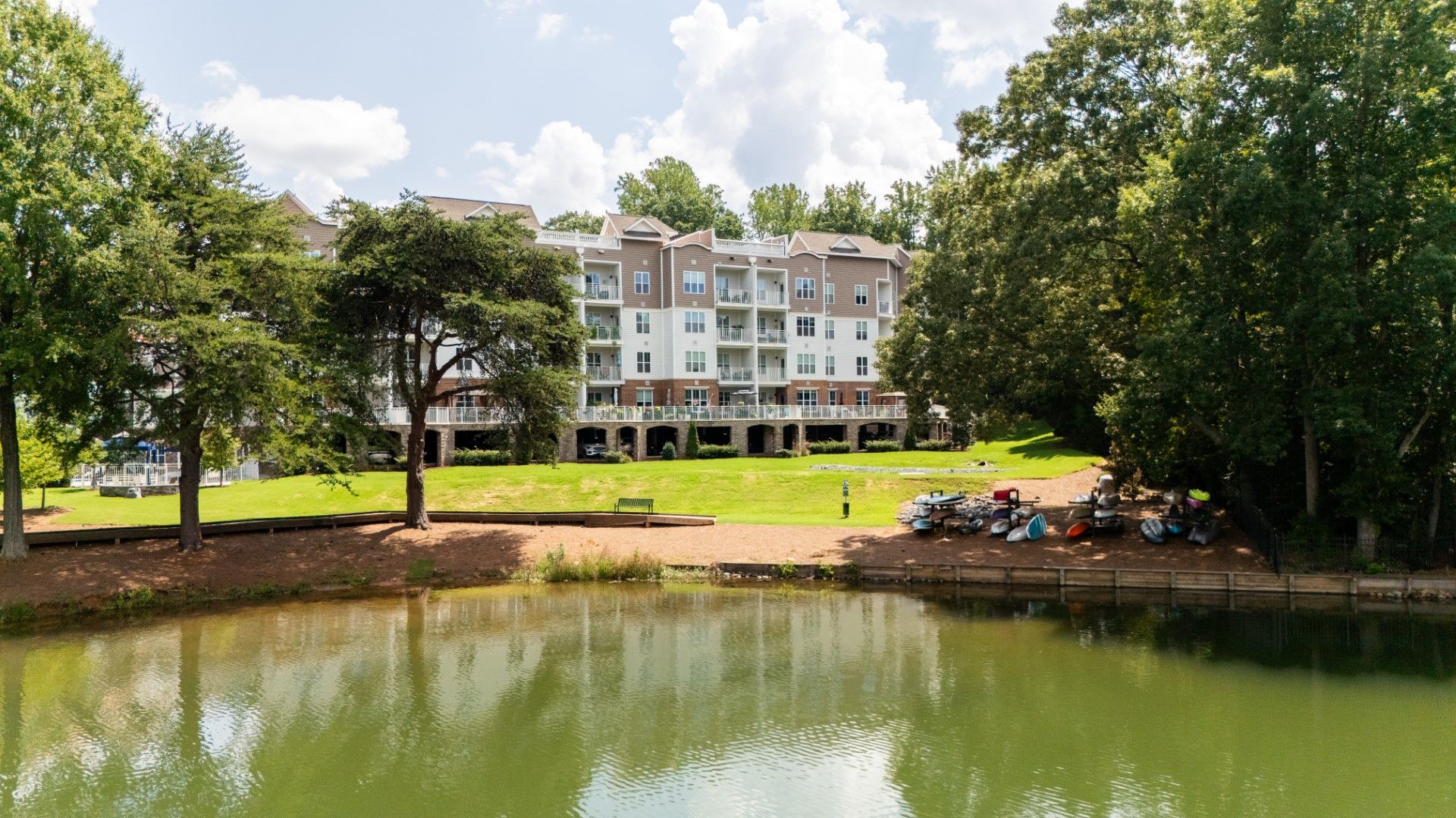 Apartment building across a pond with trees and a grassy bank under a partly cloudy sky