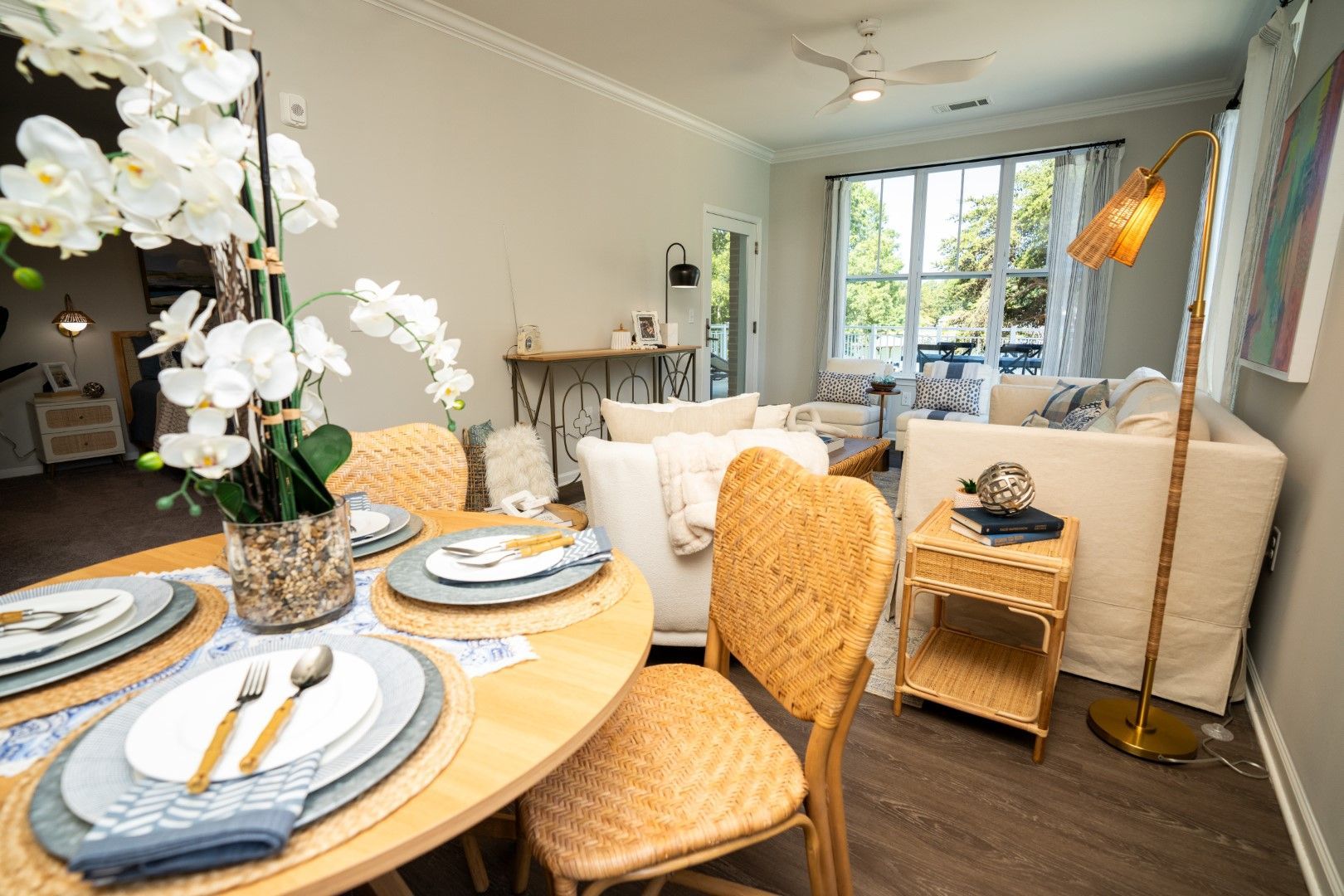Dining area with a round table set for a meal, wicker chairs, and a living room in the background with a large window at The Osprey at Lake Norman, offering luxury apartments in Mooresville, NC.