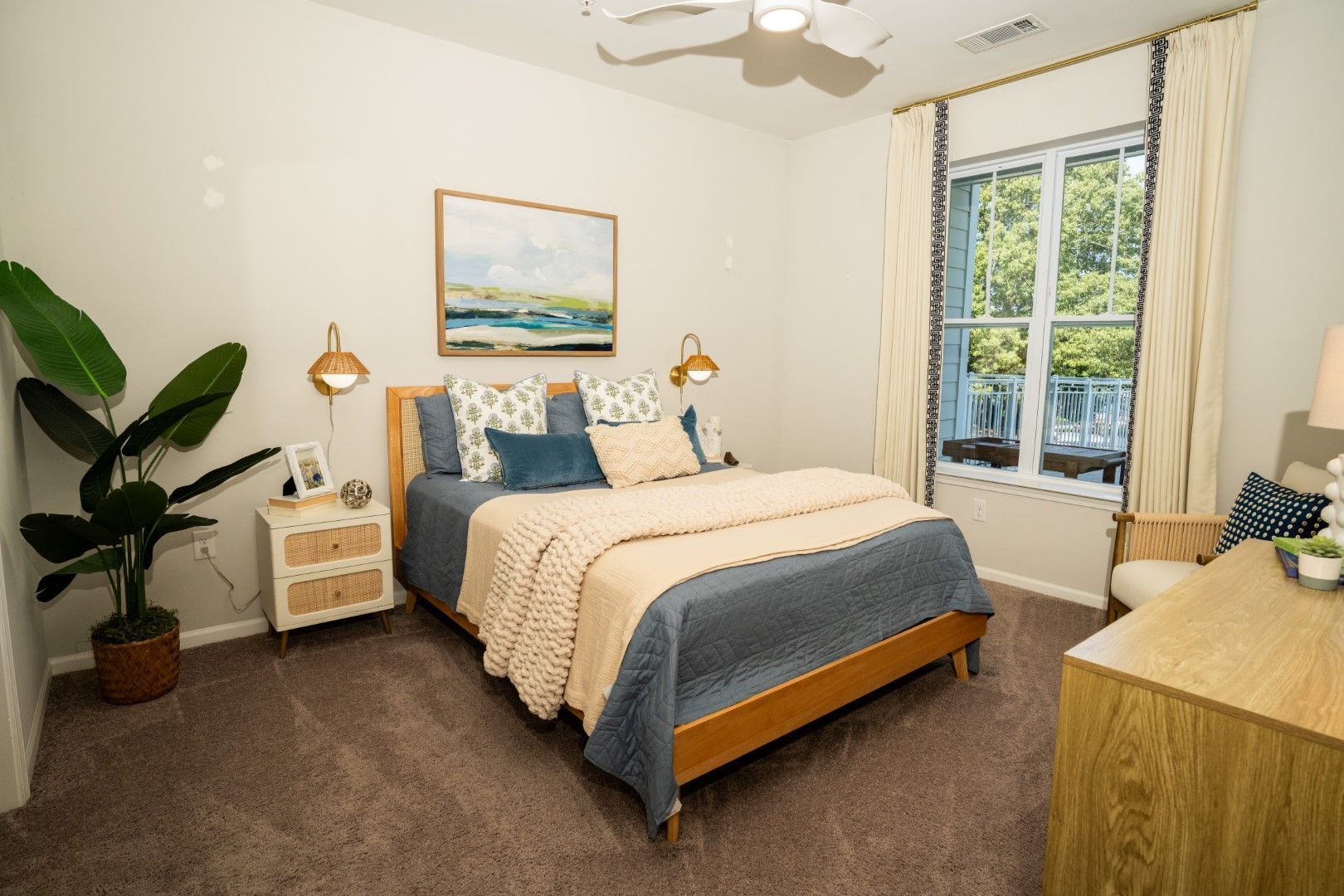 Bedroom with bed, nightstand, dresser, large plant, window with curtains, and artwork at The Osprey at Lake Norman, offering Mooresville apartments for rent.