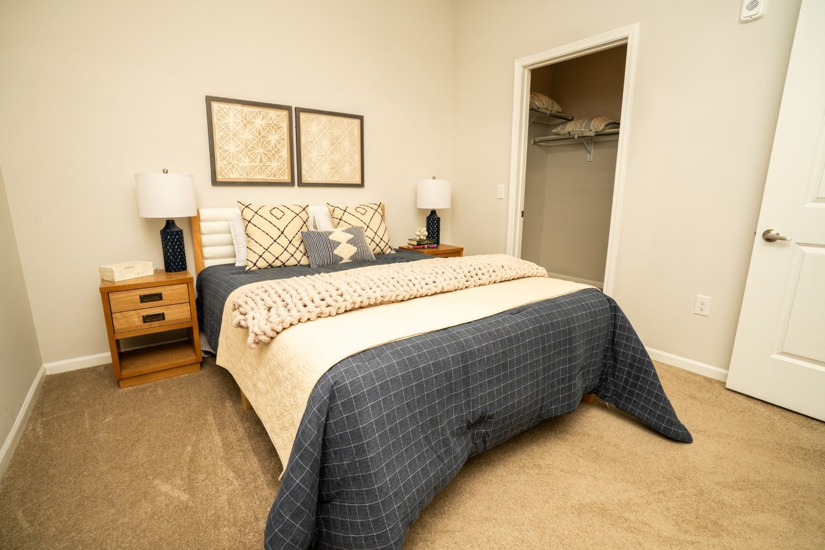 Bedroom with bed, nightstands, lamps, artwork, closet, and neutral-toned carpet and walls at The Osprey at Lake Norman, offering Mooresville apartments for rent.