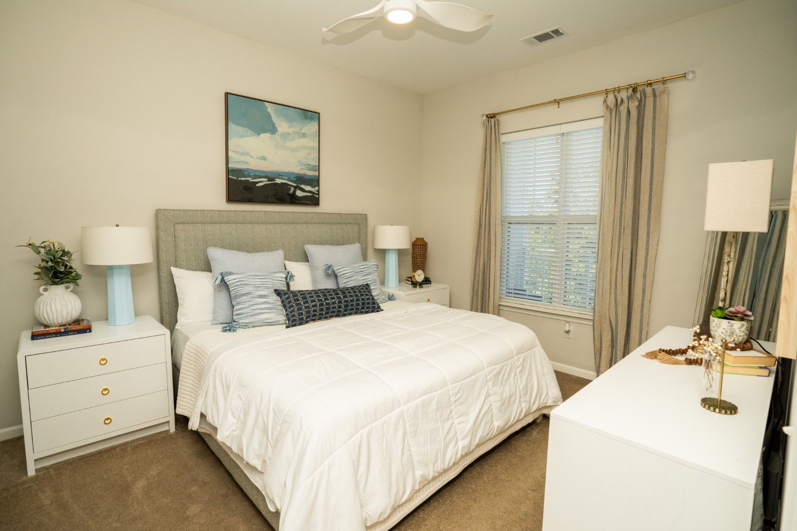 Bedroom with a bed, nightstands, dresser, artwork, and window with curtains; neutral tones at The Osprey at Lake Norman, offering Mooresville apartments.