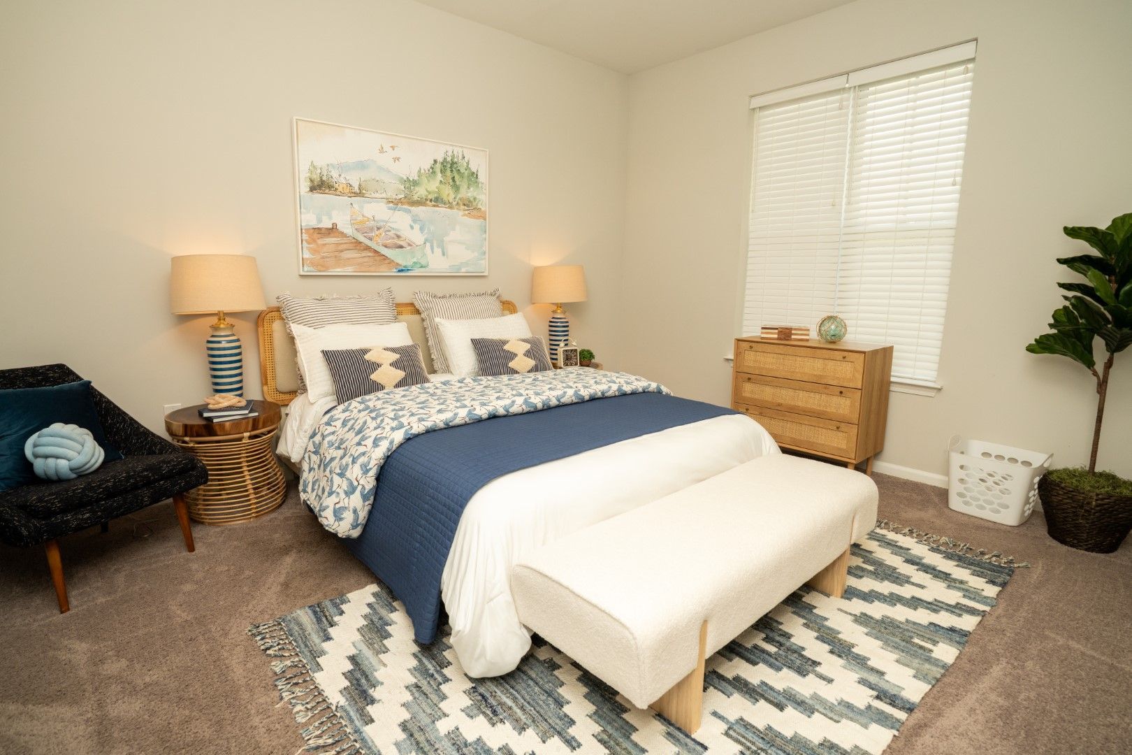 Bedroom with blue and white bedding, art, a wooden dresser, and a large plant at The Osprey at Lake Norman, offering luxury apartments in Mooresville, NC.