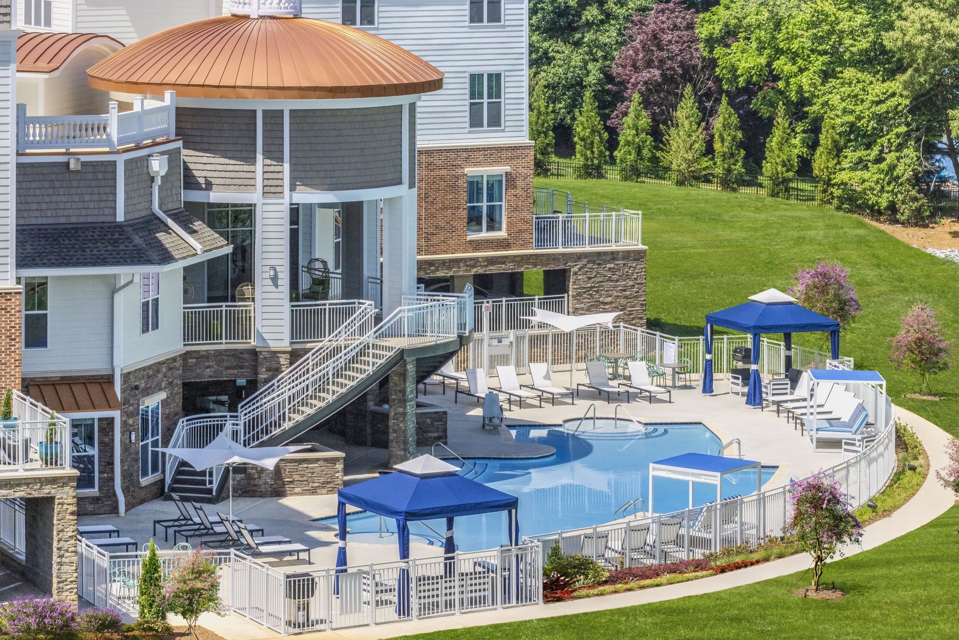 Outdoor pool with blue umbrellas and lounge chairs beside a multi-story building and grassy hillside
