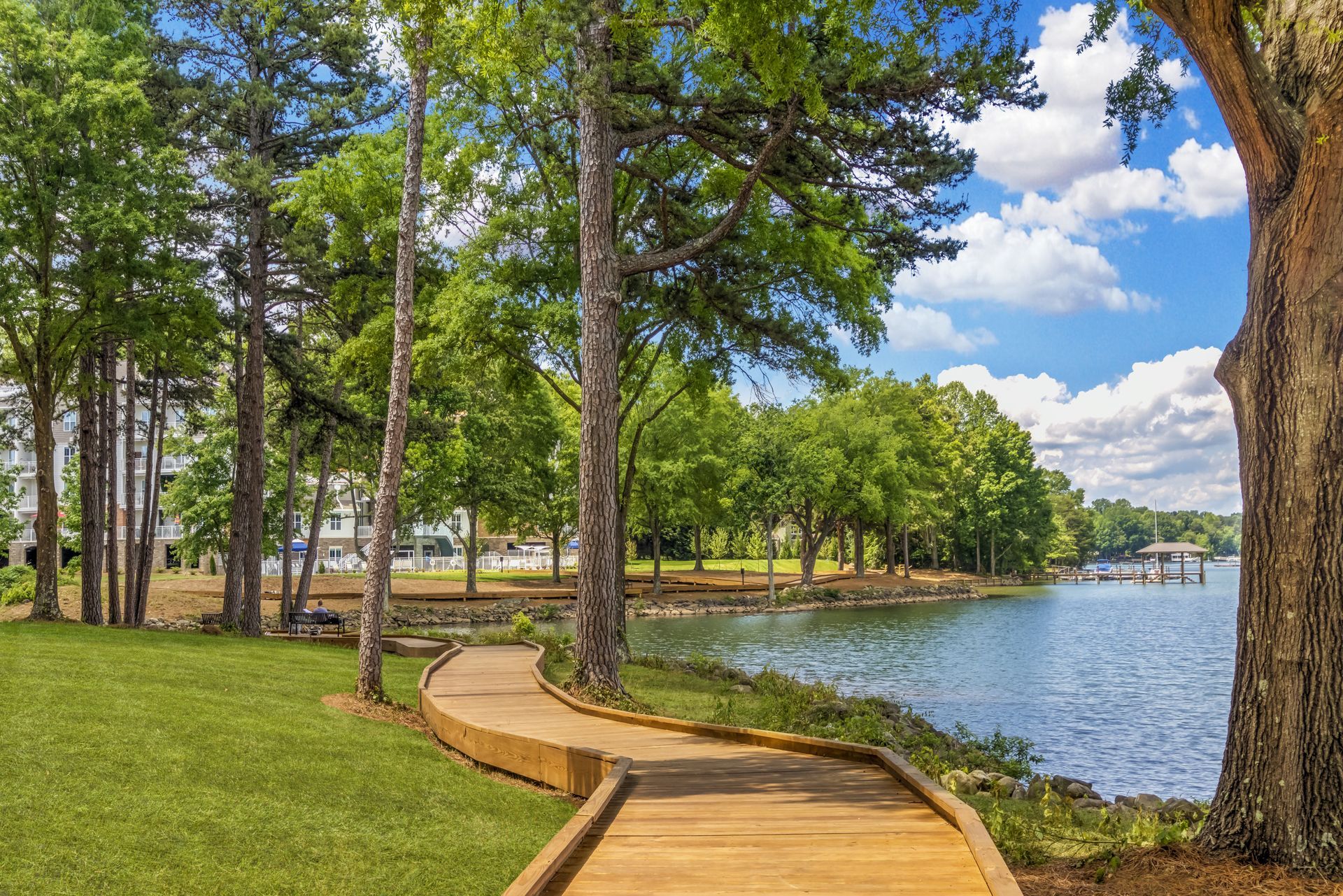 Wooden boardwalk along a tree-lined lakeshore under a blue sky with clouds