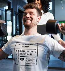 Man smiling while lifting dumbbells in a gym, with a trainer standing behind him