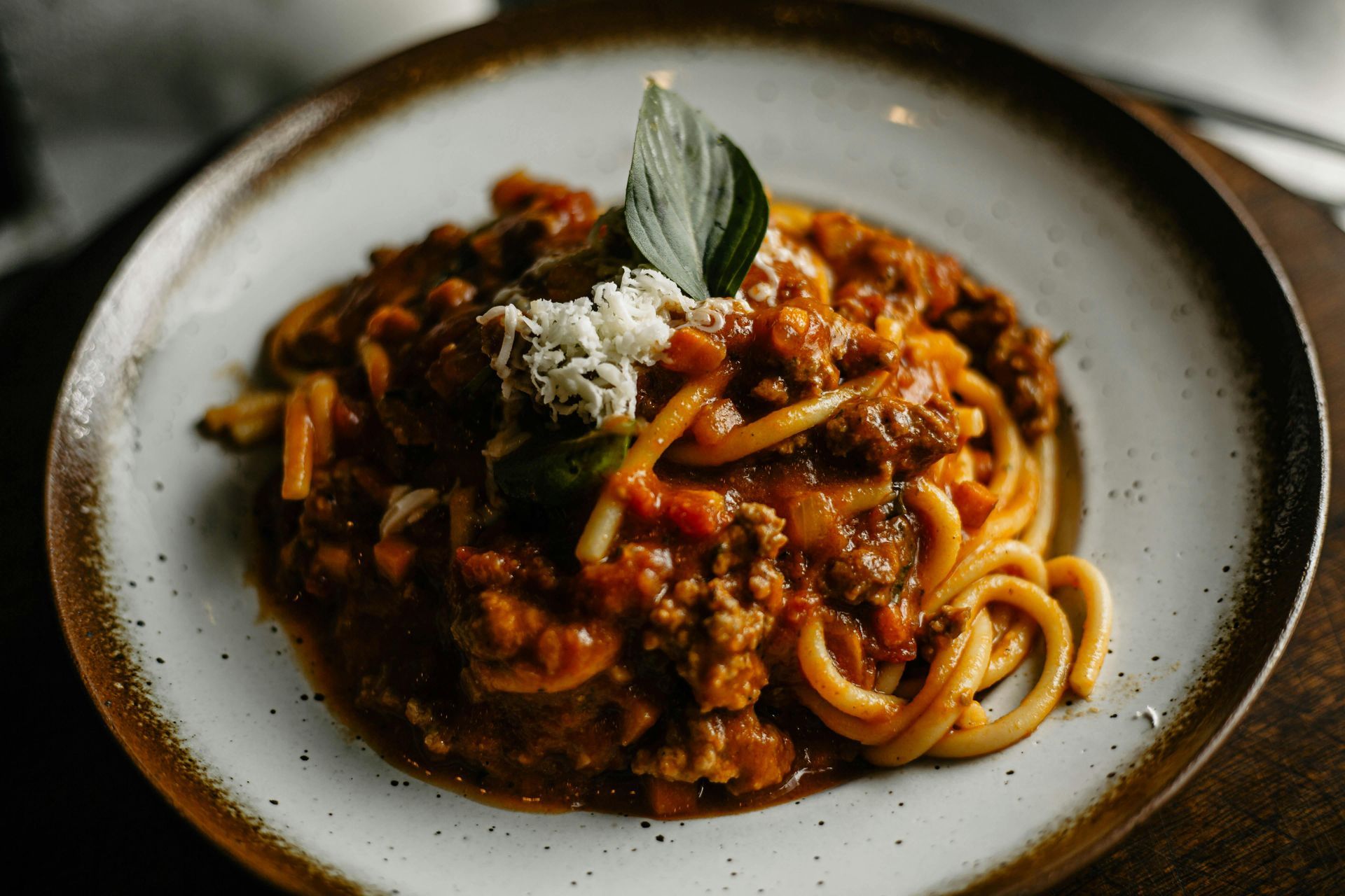 Pasta in rich tomato meat sauce on a white plate, topped with grated cheese and a basil leaf