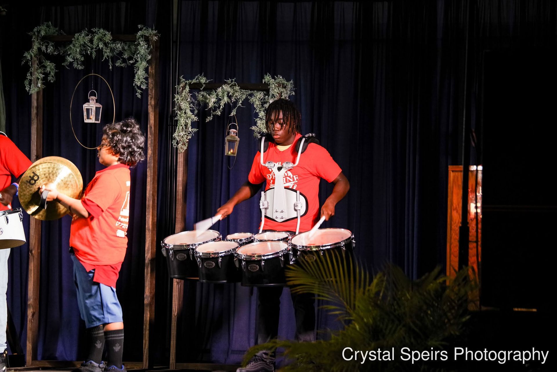 Drummers performing onstage under hanging greenery, one playing timpani and another at a large brass instrument