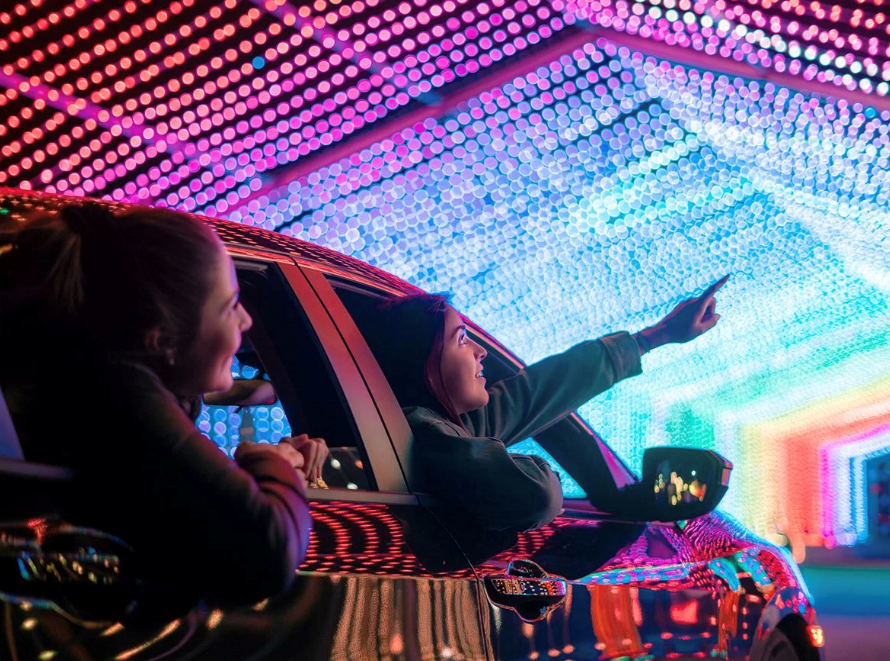 People in a car under a glowing pink and blue LED tunnel, one pointing ahead