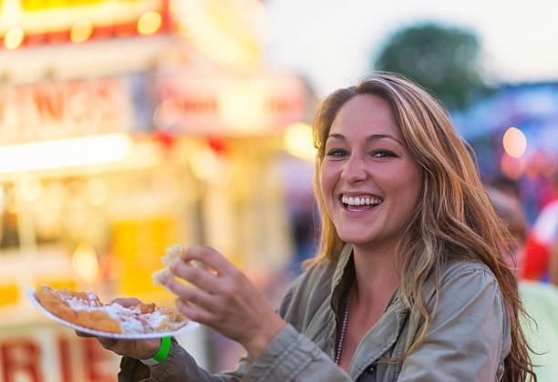Smiling woman holding a plate of food at a festive outdoor fair with bright lights in the background