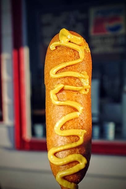 Corn dog topped with mustard on a stick, against a blurred restaurant window background