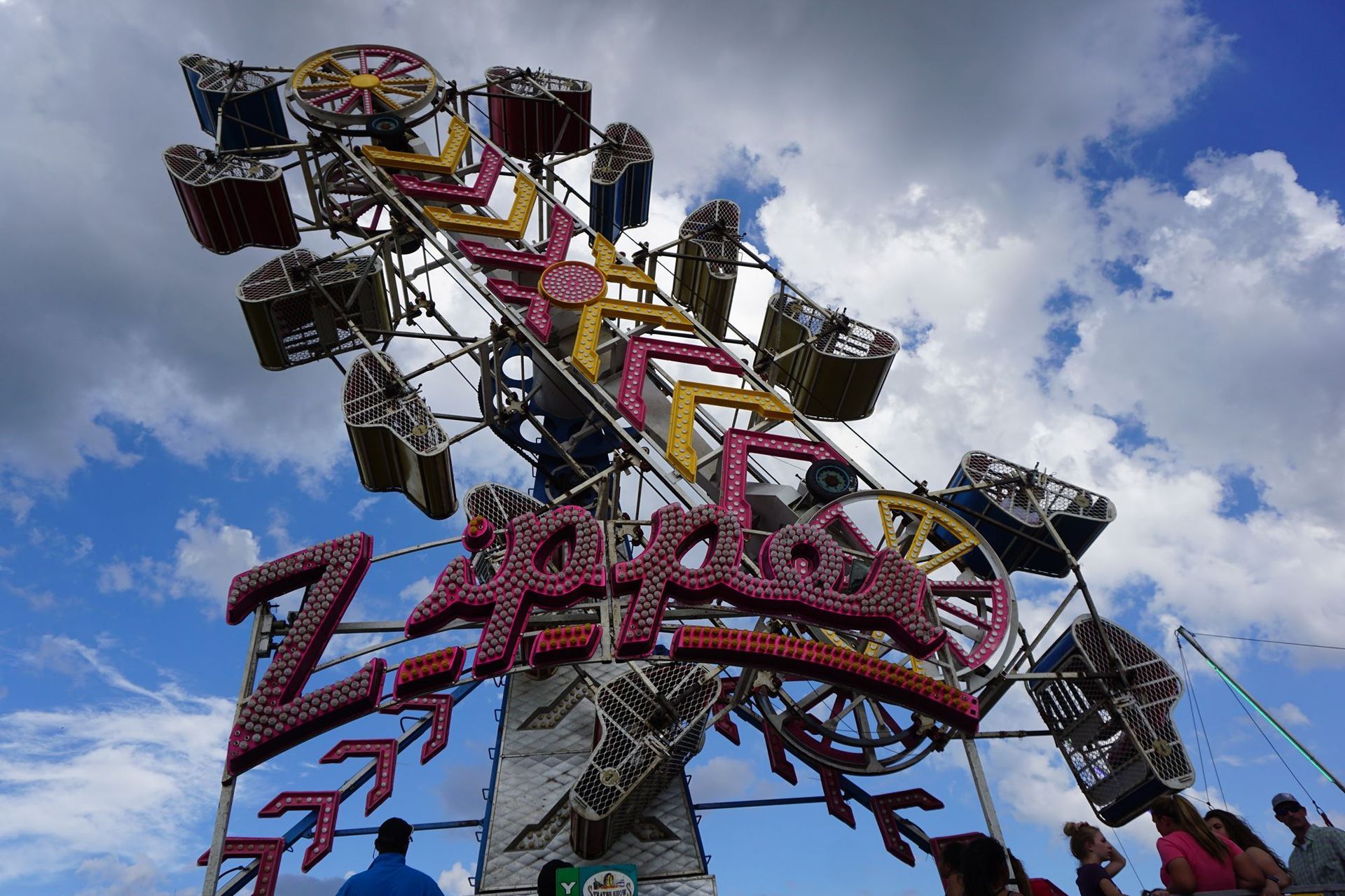 A colorful carnival ride with gondolas and large pink lettering against a cloudy sky, seen from below.