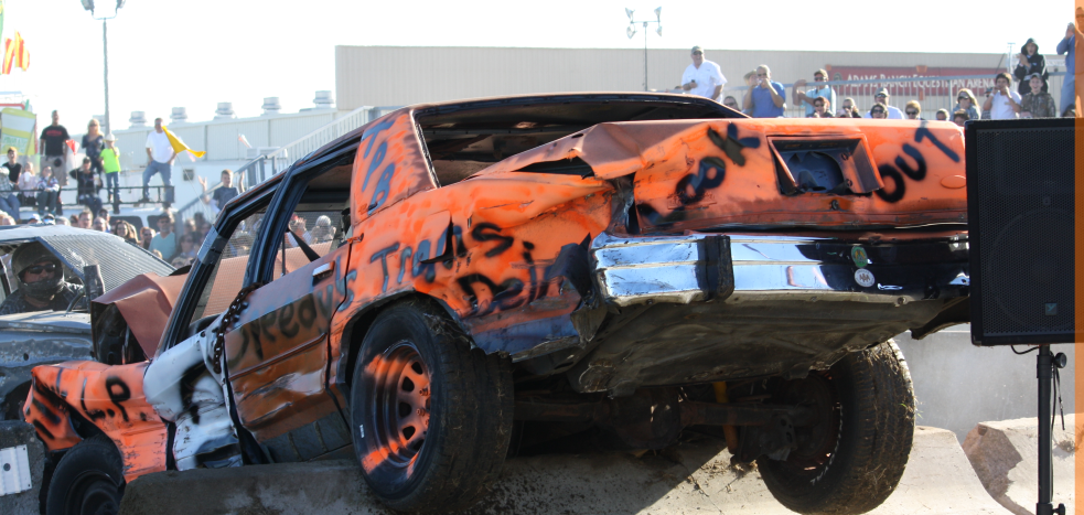 Orange race car mid-crash with damaged body, surrounded by smoke at a demolition derby track