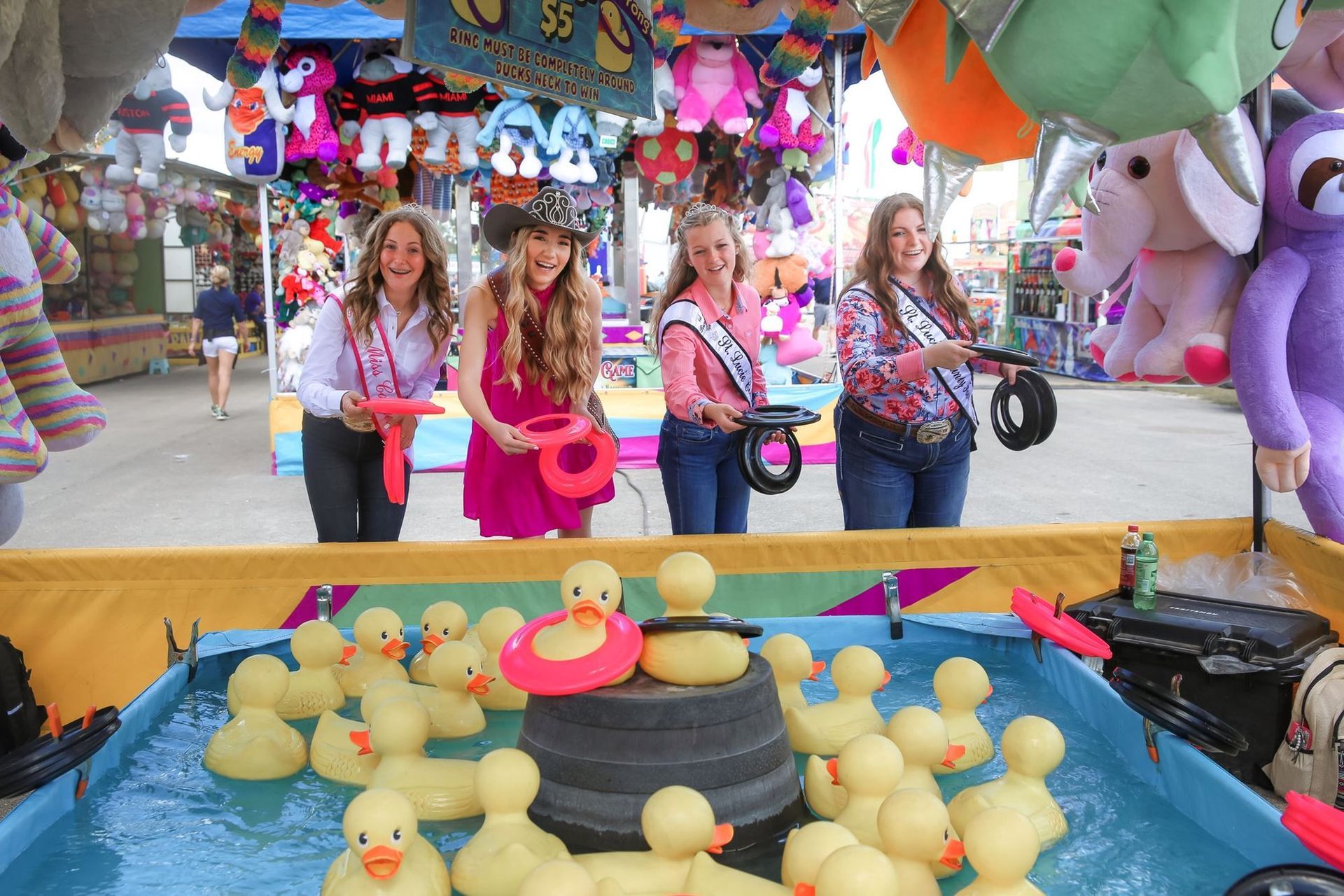 Women in pink shirts at a carnival game, aiming darts at rubber ducks in a colorful booth.