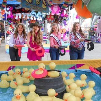 Four women at a carnival duck ring toss game, holding pink rings in front of rubber ducks and tires