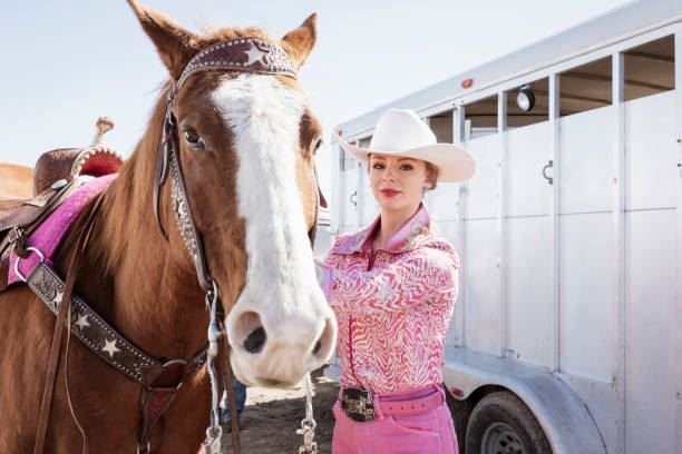 Woman in pink outfit and white hat standing beside a saddled brown horse near trailers