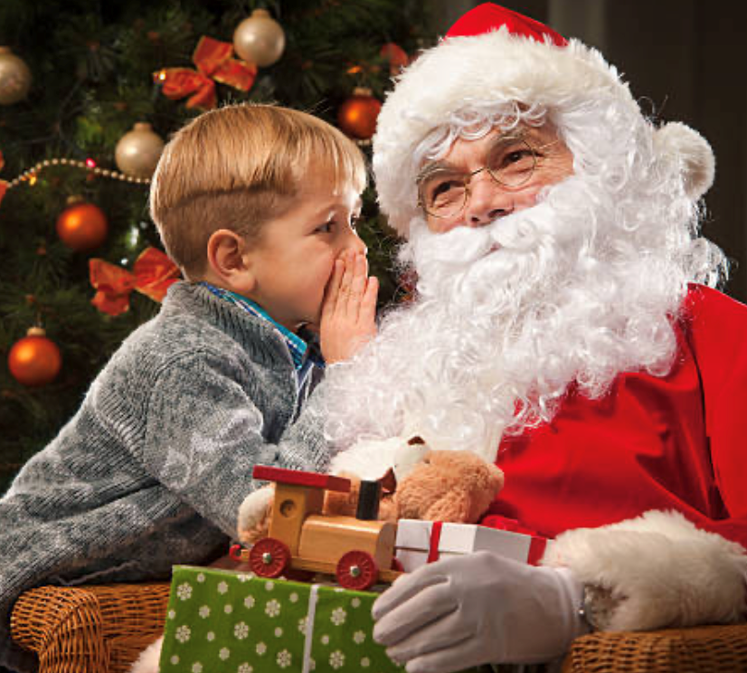 Child whispering to Santa beside a Christmas tree, holding a toy train and gift box.