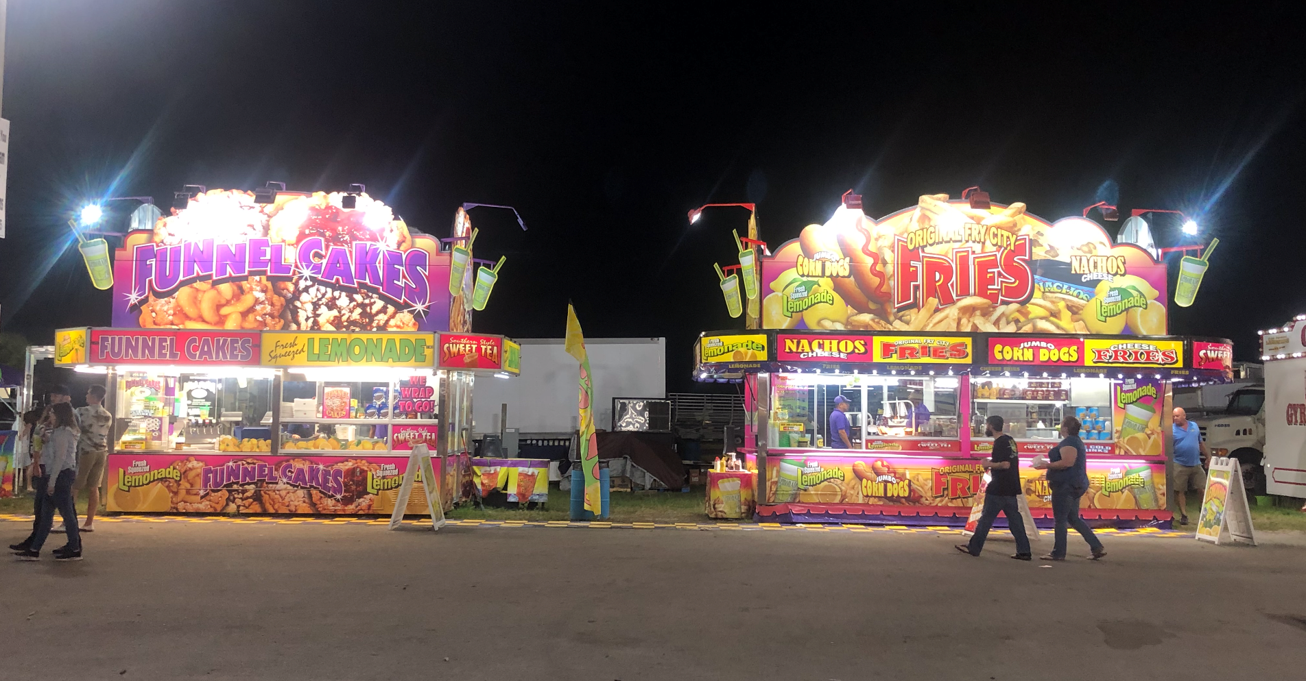 Two brightly lit carnival food stalls at night, with people walking in front of them.