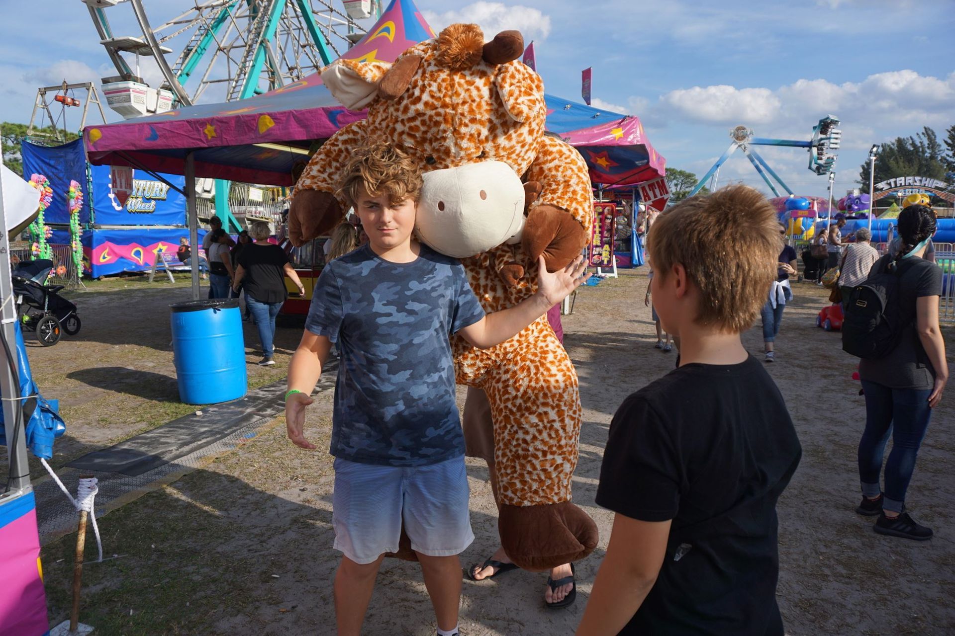 Two children posing with a giraffe mascot at a colorful outdoor fairground.