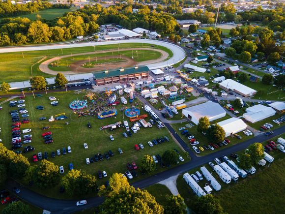 Aerial view of a small outdoor fair with tents, parked vehicles, and a dirt racetrack at sunset