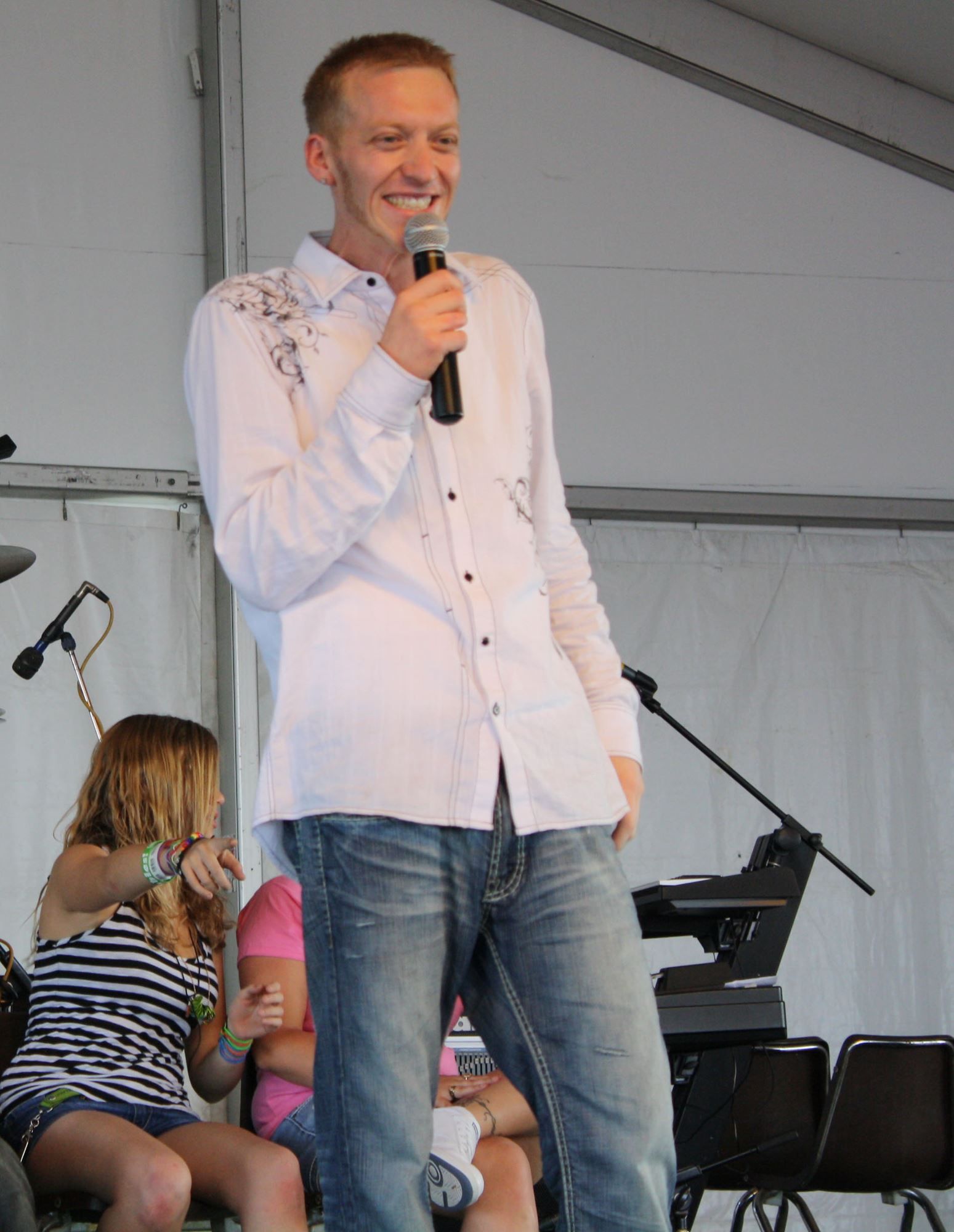 Man speaking into a microphone on an outdoor stage, with seated people and musical equipment behind him.