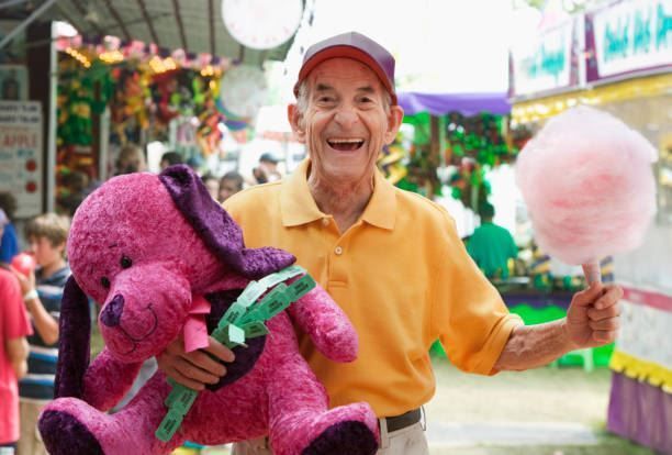 Smiling man in yellow shirt holding a purple stuffed toy at a colorful carnival booth