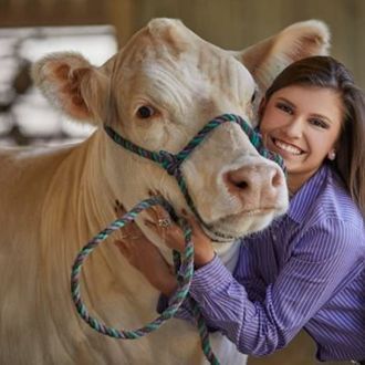 Smiling woman in a purple shirt hugging a light-colored cow with a rope halter in a barn.