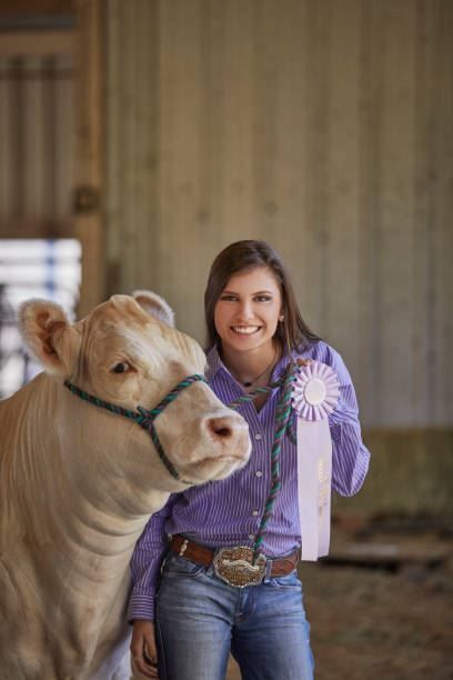 Woman beside a tan cow, holding a purple ribbon in a barn.