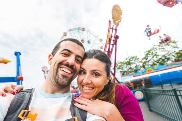 Smiling couple taking a selfie at an amusement park with a Ferris wheel and colorful rides behind them