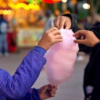 Hands holding pink cotton candy at a fair, with a blurred colorful background