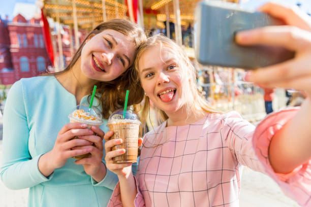 Two friends taking a selfie and holding iced drinks on a sunny street