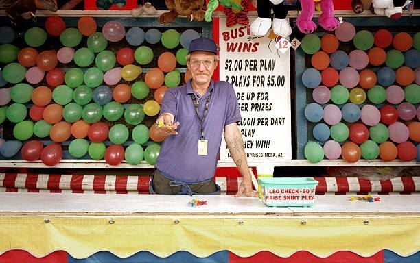 Man in purple shirt standing at a carnival booth with colorful balloons and a sign behind him