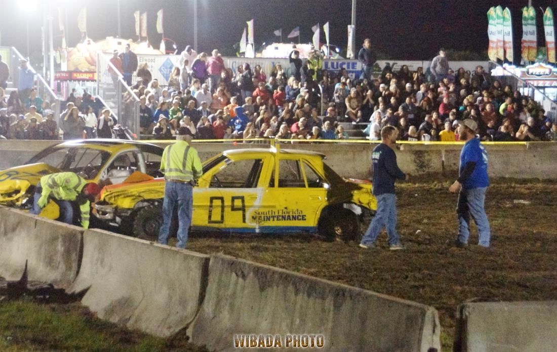 Yellow race car with crew on a dirt track at night, crowd watching from the stands