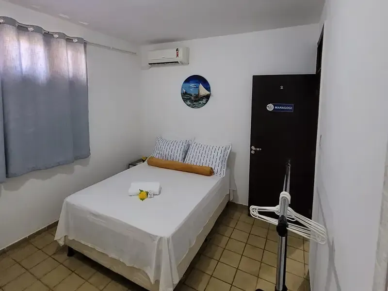 Quarto pequeno com uma cama, uma janela com cortinas e uma porta escura com um ar condicionado em cima.