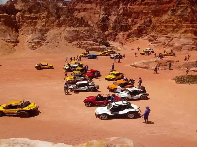 Um grupo de buggies em uma praia de areia vermelha, com pessoas e um penhasco vermelho-acastanhado ao fundo.