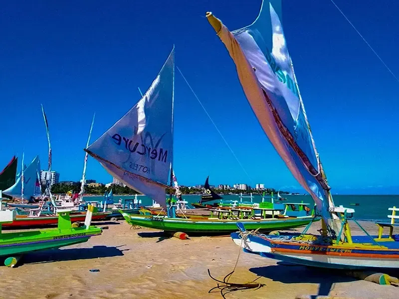 Barcos à vela coloridos em uma praia arenosa sob um céu azul brilhante, perto do oceano.