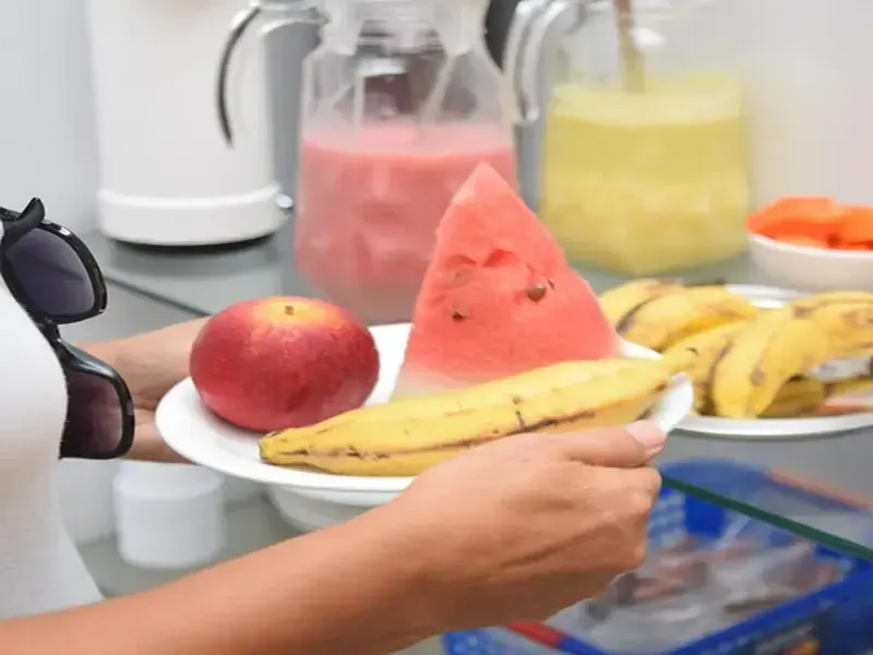Mulher segurando um prato de frutas: maçã, melancia e banana. Sucos e outras frutas ao fundo.