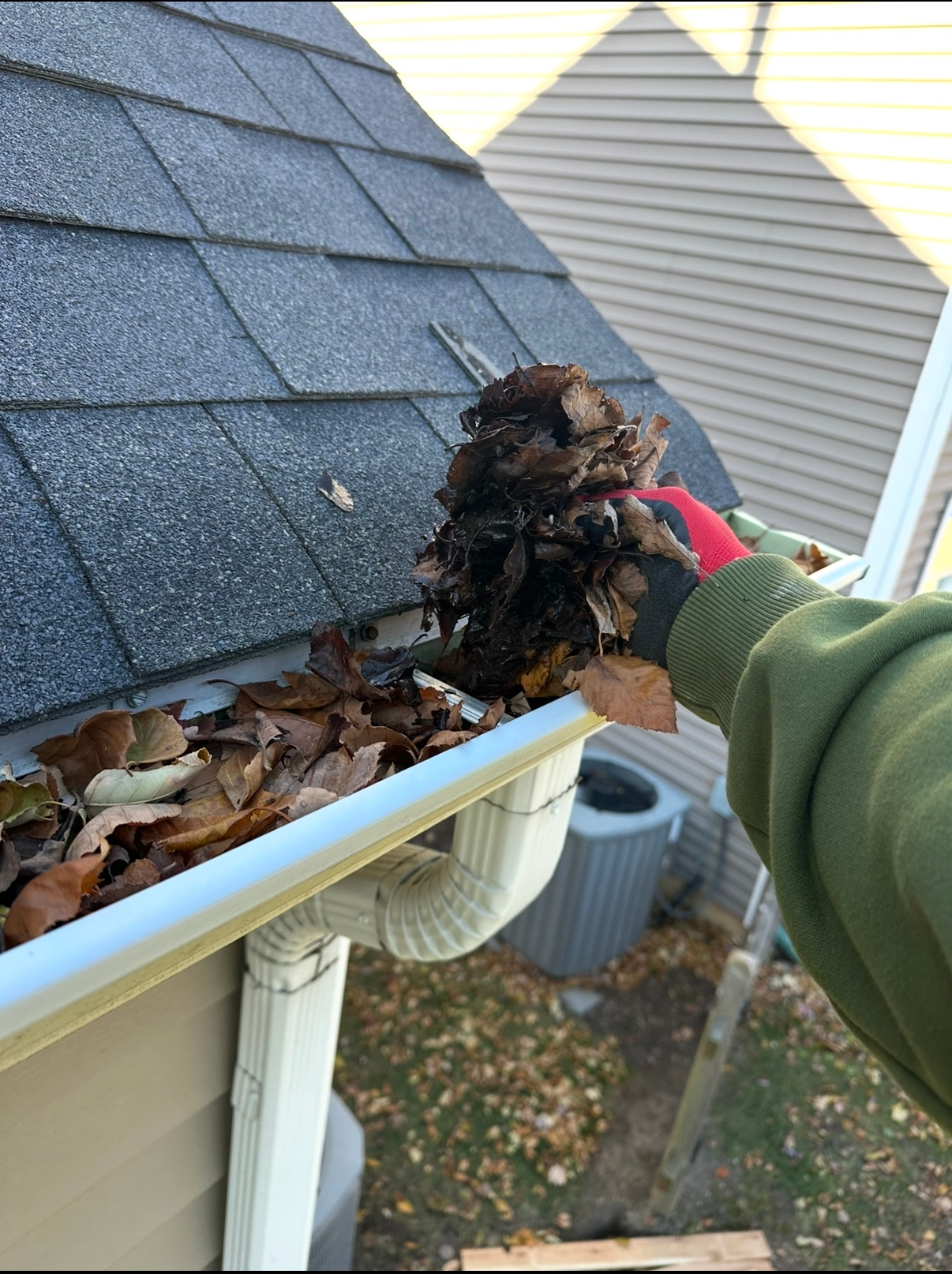 A person wearing a green sweatshirt and red-trimmed gloves pulls a clump of wet leaves out of a white house gutter.