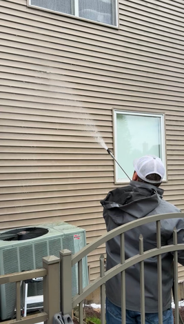 A person in a gray jacket and white hat uses a pressure washer to clean tan horizontal siding on a house.
