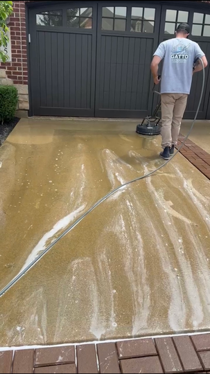 A person in a high-visibility orange jacket uses a pressure washer to clean a concrete sidewalk outside a shopping plaza.