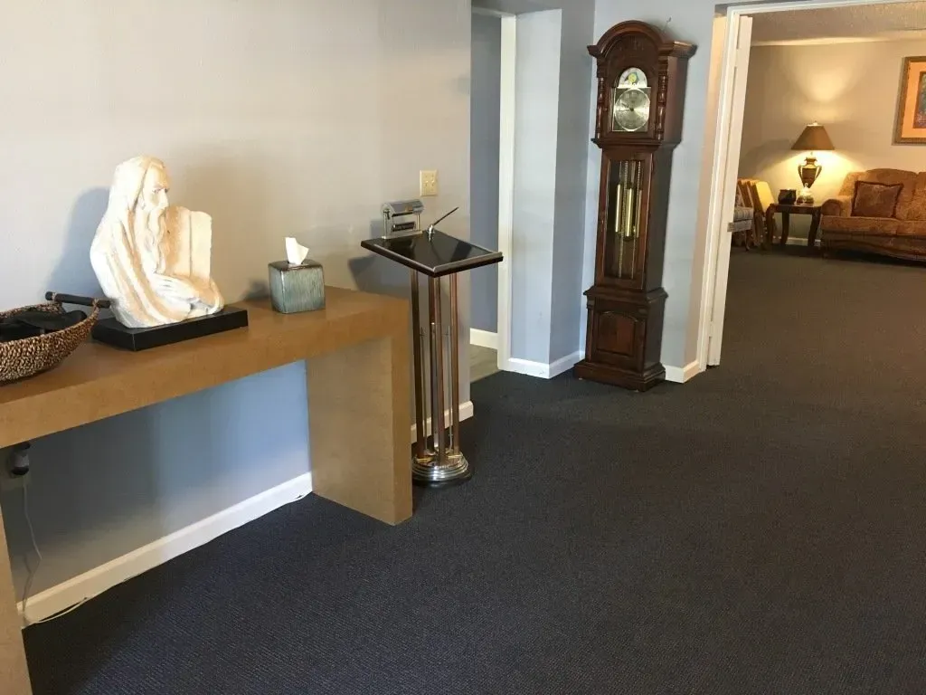 Hallway with wooden console table, white statue, grandfather clock, and a lit room in the background