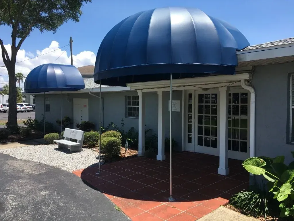 Blue awnings over a white building entrance with a glass door, red brick patio, and landscaped shrubs