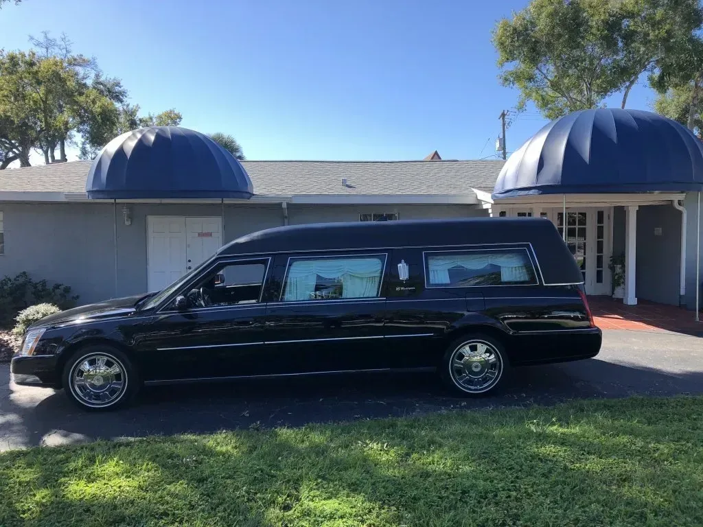Black hearse parked on grass in front of a building with blue awnings.