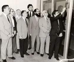 Men in suits gather indoors, one pointing at a display board while others watch attentively.
